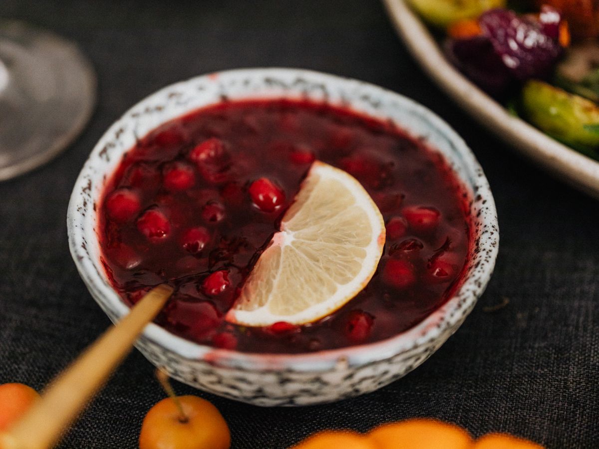 A small bowl of cranberry sauce with whole berries and a lemon slice, roasted vegetables behind it