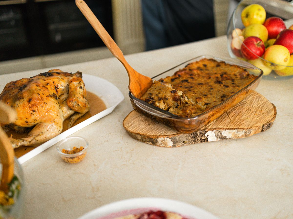 Baked bread stuffing in a glass dish with a wooden spoon, a roast bird on a platter beside it