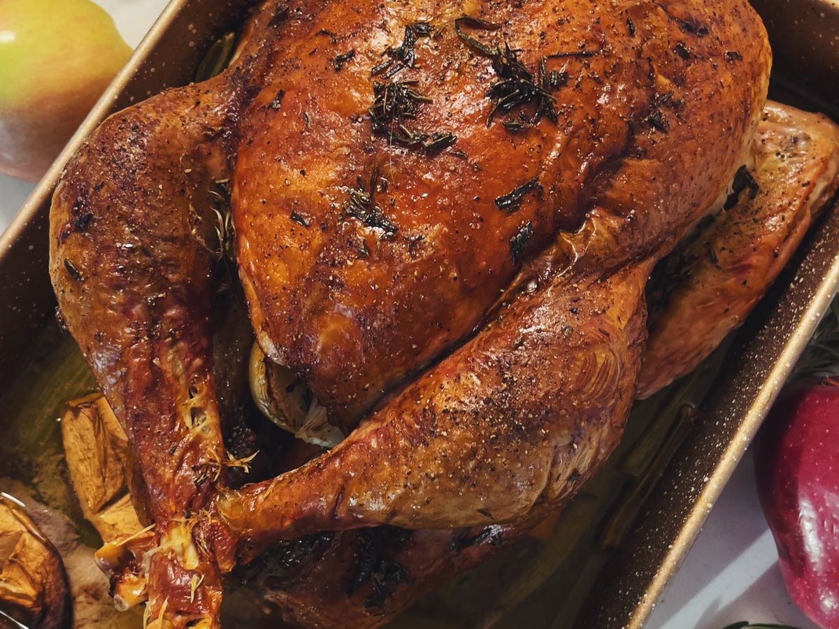 A whole roast turkey with browned skin and herbs in a roasting tin, apples beside it